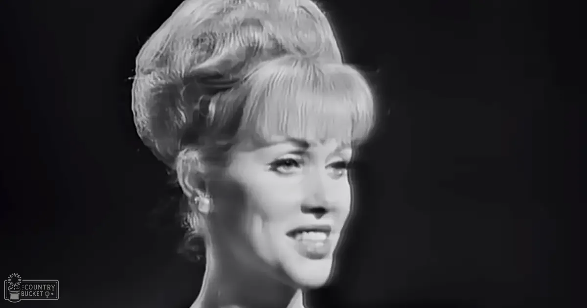 A woman, likely an adult female, dressed in a light-colored, mid-20th-century-style dress with short sleeves and high neckline, wearing dangling earrings against a plain black background.
