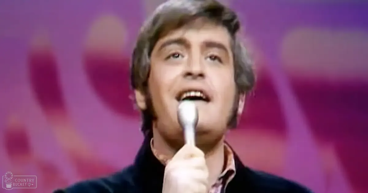 An adult male singer wearing a dark jacket over a collared shirt with a floral or paisley pattern, holding a microphone on stage.
