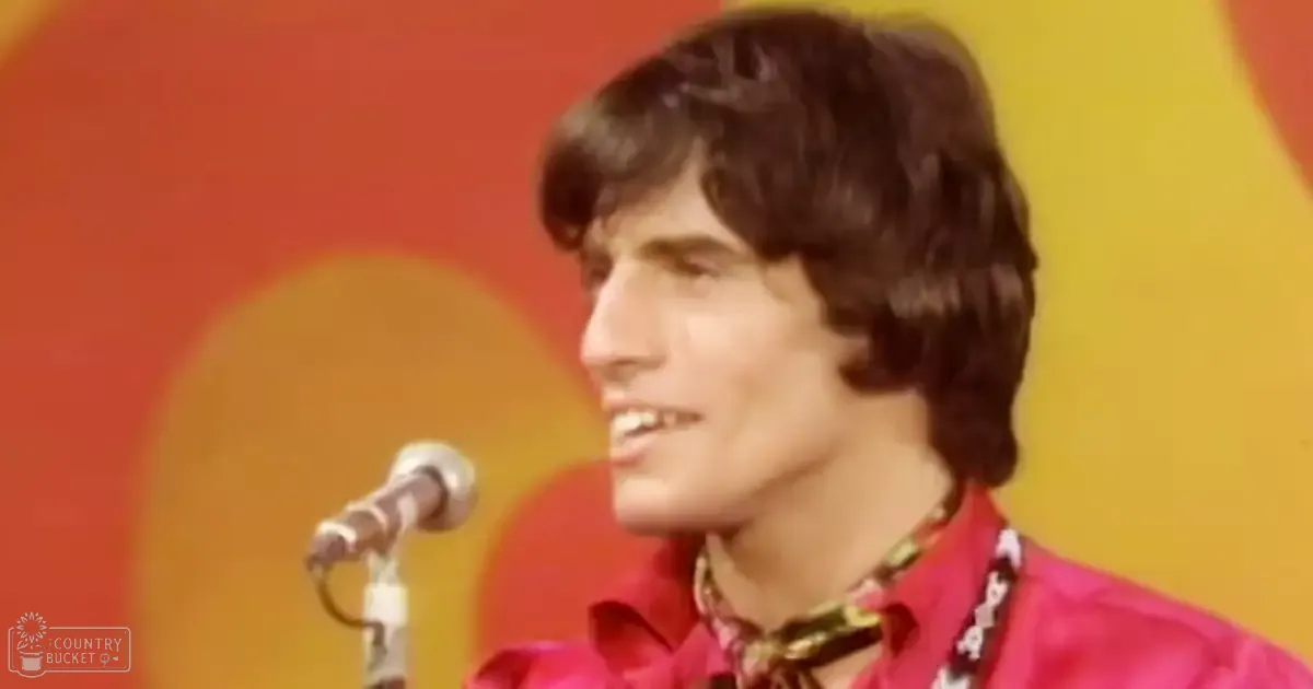 A young adult male wearing a red shirt with a patterned tie, standing in an indoor setting with stage-like elements including a microphone stand and colorful backdrop.
