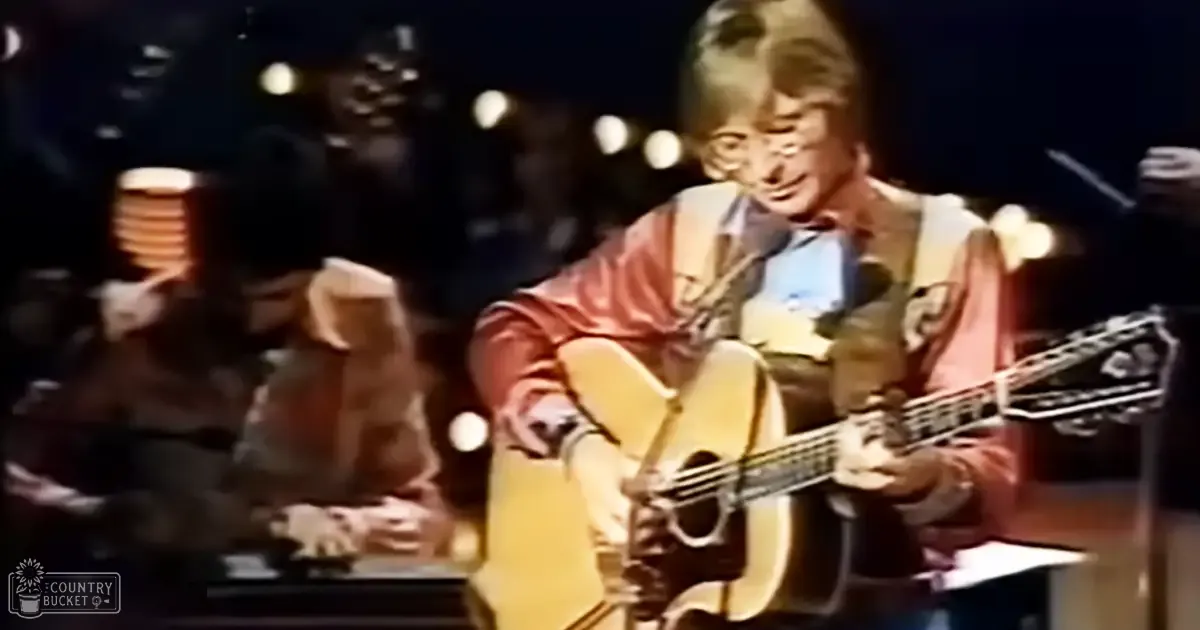 An adult male musician wearing a red jacket over a blue patterned shirt performs on stage under artificial lighting, surrounded by concert equipment and an audience.