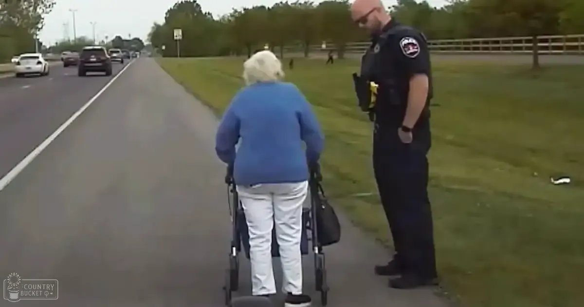 A police officer in a dark blue uniform interacts with an older adult wearing a blue sweater, white pants, and black shoes. They are on a suburban street with a grassy median and trees along the road.
