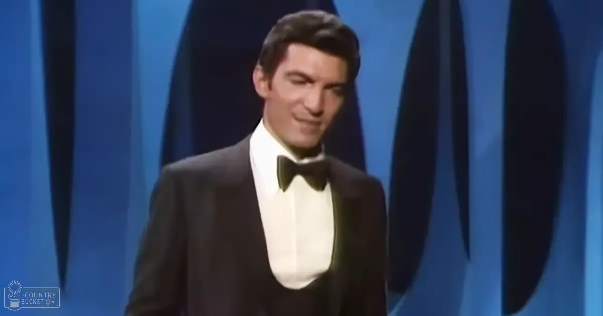 A young adult male in a black tuxedo with a white dress shirt and a black bow tie, standing against a blue curtain backdrop at an event setting.