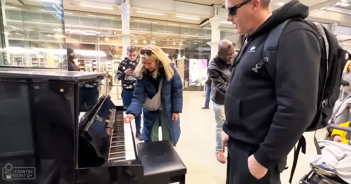 An adult female plays an upright piano in a public space, possibly a train station or shopping mall; she wears a dark blue puffer jacket and green pants.