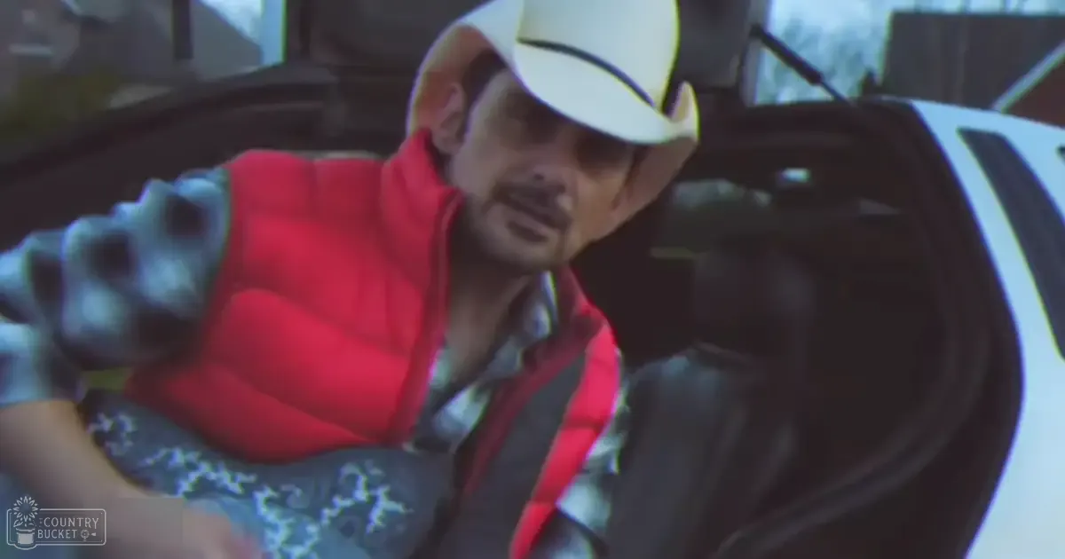A middle-aged man wearing a white baseball cap, red puffer vest over a geometric-patterned garment, dark pants, stands near an open vehicle door in a rural or suburban outdoor setting with some greenery visible in the background during daytime.