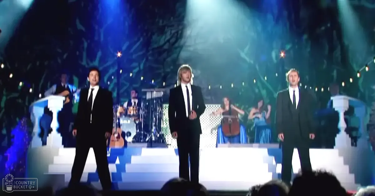 Adult males in dark suits perform at an indoor concert venue with a forest-like backdrop and blue-green lighting effects.