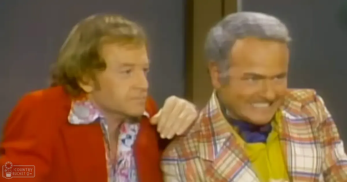 A mid-aged male wearing a red patterned jacket and another man with an orange, white, blue, and red striped plaid shirt in what appears to be a simple indoor studio setting with neutral walls.