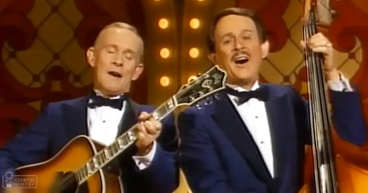 A middle-aged male singer wearing a dark blue tuxedo and holding an acoustic guitar, performing with another middle-aged male singer on stage against a patterned backdrop with warm tones.