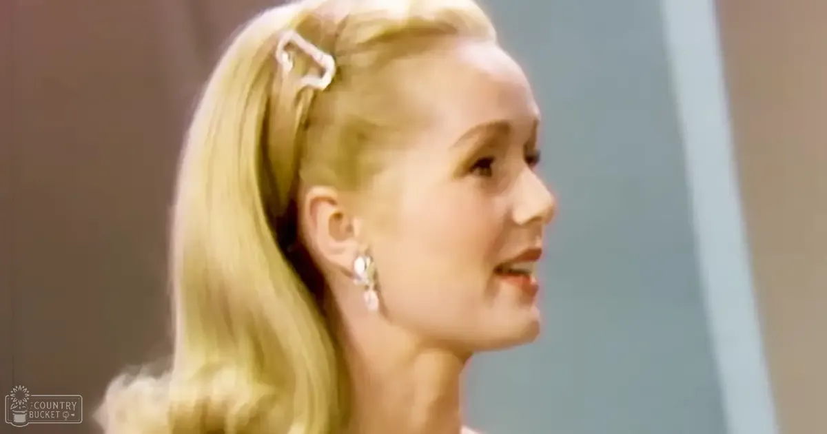 A young adult female with long blonde hair styled with a barrette, wearing earrings and vintage-style attire indoors, well-lit room during daytime.