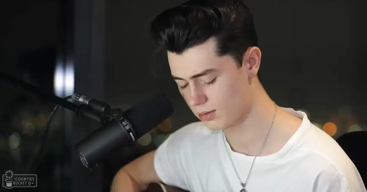 A young adult male wearing a white t-shirt with short sleeves, standing indoors near recording equipment under artificial lighting at night or evening.