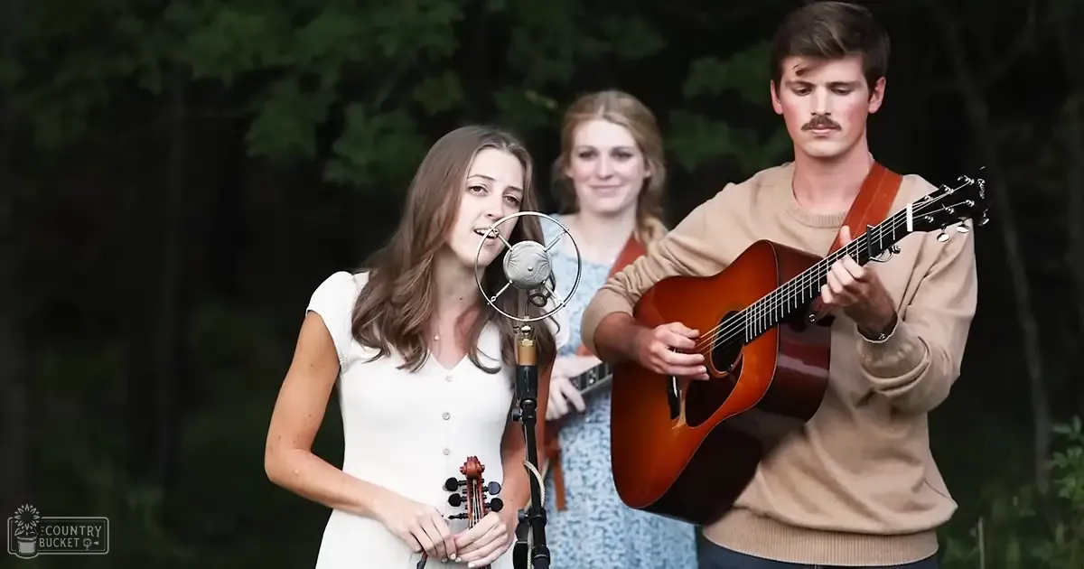 A young adult female singer in a white sleeveless dress performs outdoors with an acoustic guitarist.