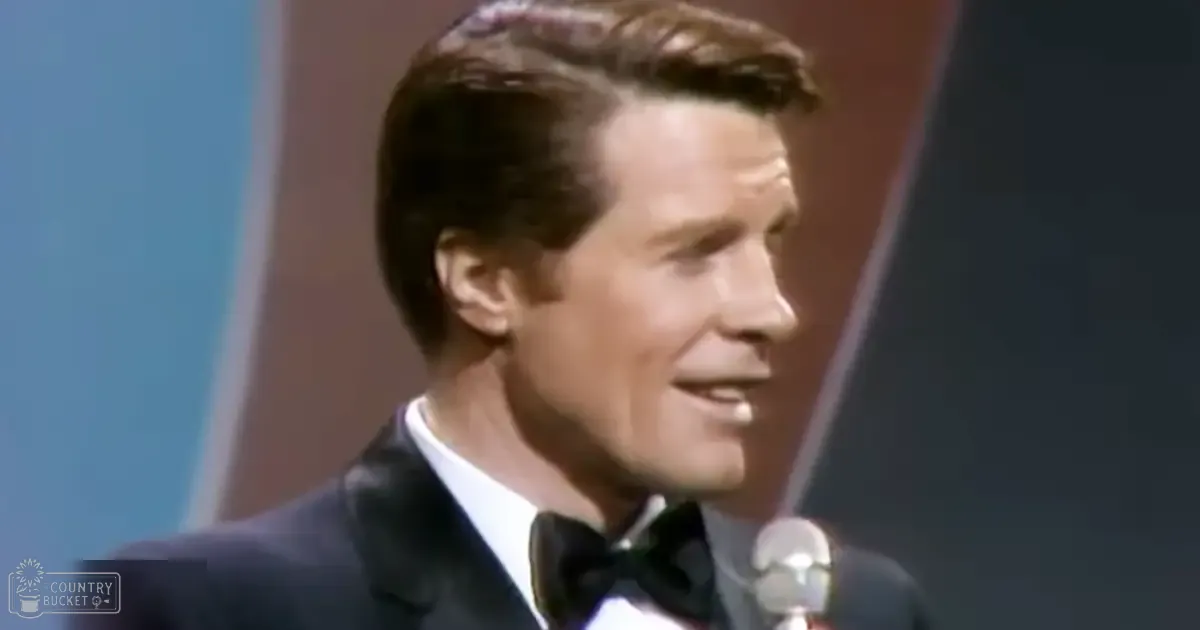 A middle-aged man in a dark tuxedo with a white dress shirt and black bow tie performs at what seems to be a formal event, with a microphone prominently featured in front of him. The background includes shades of blue and brown, suggesting an indoor setting possibly designed for performances or speeches.