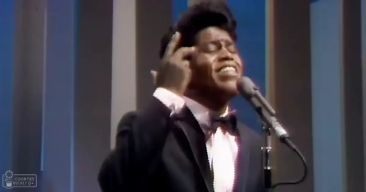 A young adult male singer in a dark suit, white shirt, and black bow tie performs into a microphone on stage under professional lighting.