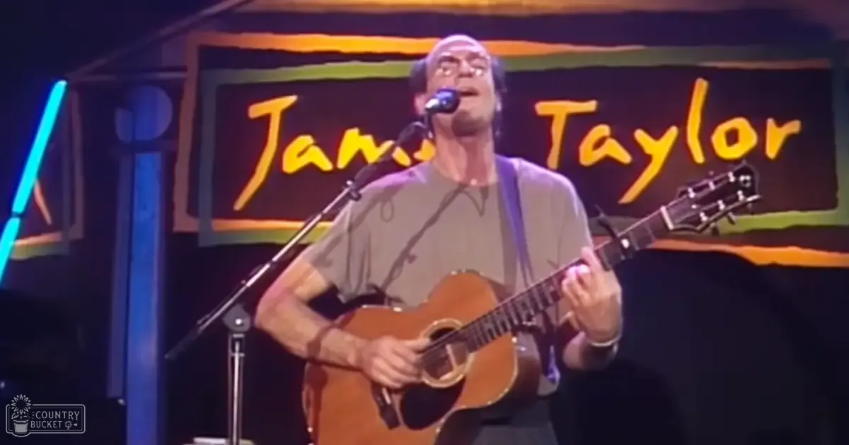 A middle-aged male musician performing at an indoor venue named 'Jams Taylor', wearing a light-colored button-up shirt with rolled-up sleeves, illuminated by stage lighting and dark backdrop.
