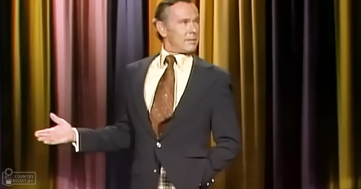 A middle-aged man wearing a dark suit with a white shirt and patterned tie performs on stage in what appears to be a comedy club, with vertical curtains of gold and brown in the background. The text 'Madly Odd' is visible in the bottom left corner.