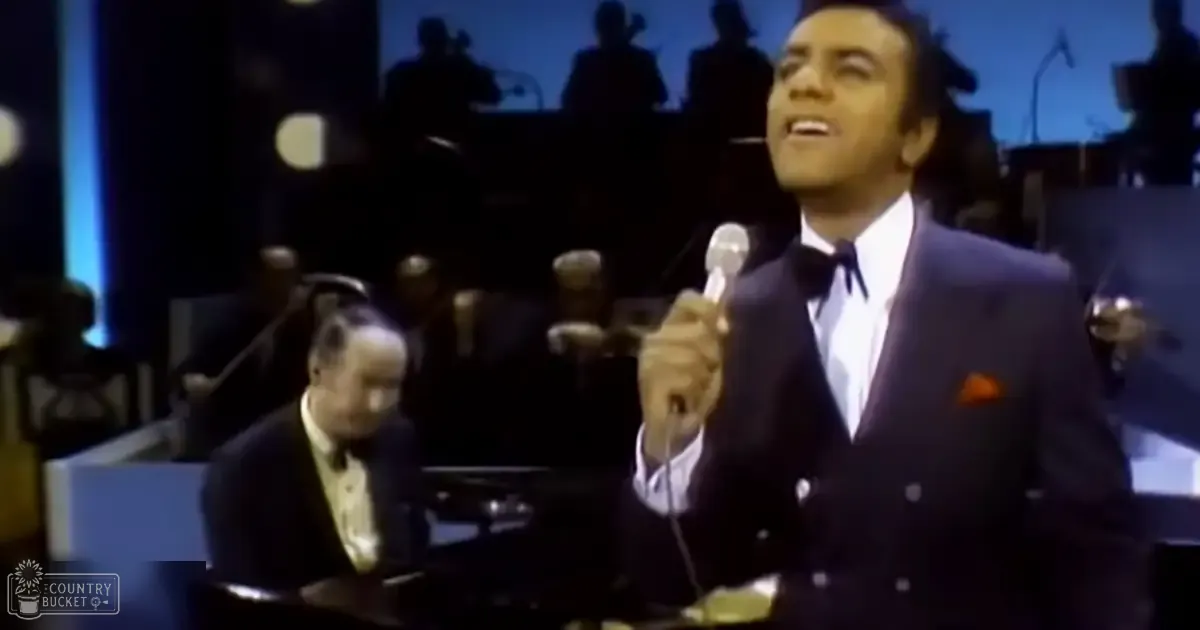 A male singer in an adult age band performs on stage with another middle-aged to older adult male seated at a piano, both dressed formally for what appears to be a live music or television production setting from the mid-20th century.
