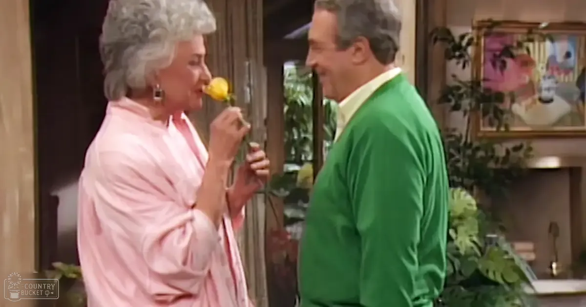An older adult female wearing a pink ruffled blouse with large hoop earrings holds a yellow flower or fruit near her mouth indoors; houseplants, framed artwork, and natural daylight suggest a cozy domestic setting.