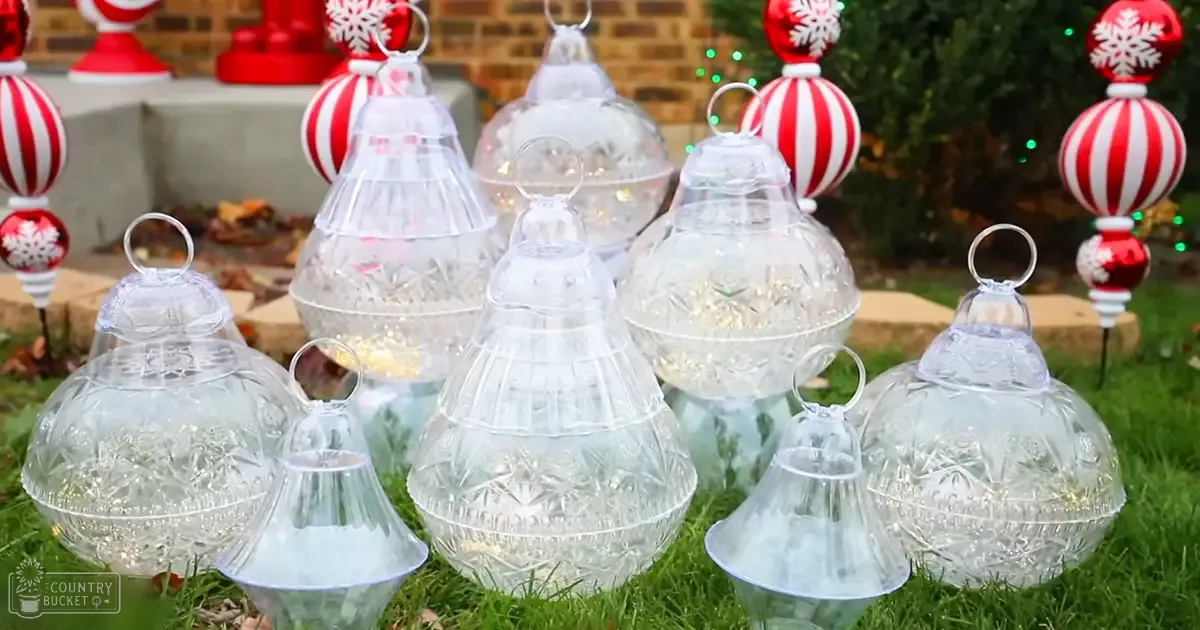 Clear glass bell-shaped ornaments with intricate designs and small lights inside them placed outdoors in a garden setting with red and white striped ornaments with snowflake patterns, brick wall, greenery, and fallen leaves indicating autumn or winter.