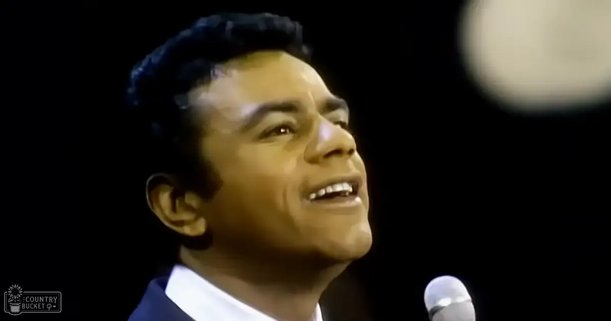 An adult male singer in a dark suit jacket singing into a microphone at an indoor performance venue with dark lighting and 'Madly Odd' text visible.