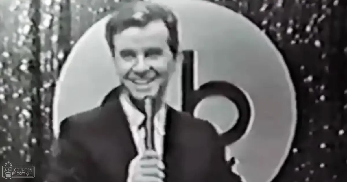 A middle-aged man in a dark suit jacket over a white shirt with a tie stands against a dark textured backdrop with the faint text 'Madly Odd', possibly indicating a performance or event context.