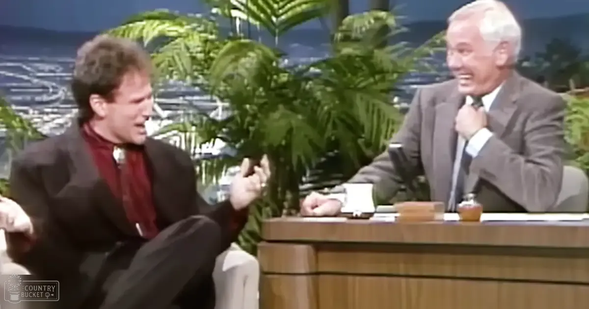 A young adult male in a dark suit jacket over a patterned red shirt, seated next to an older man in a gray suit and white shirt, both on a talk show set with tropical decor.