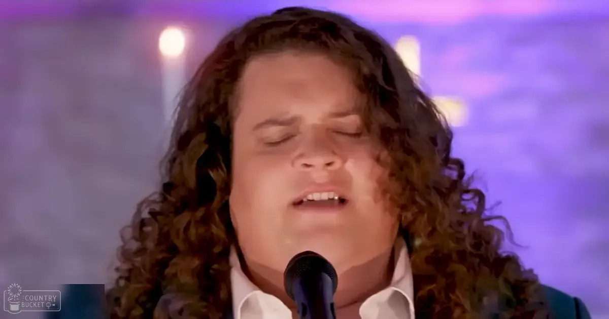 A young adult male singer in a relaxed white button-up shirt at an indoor performance venue with stage lighting, holding a microphone.