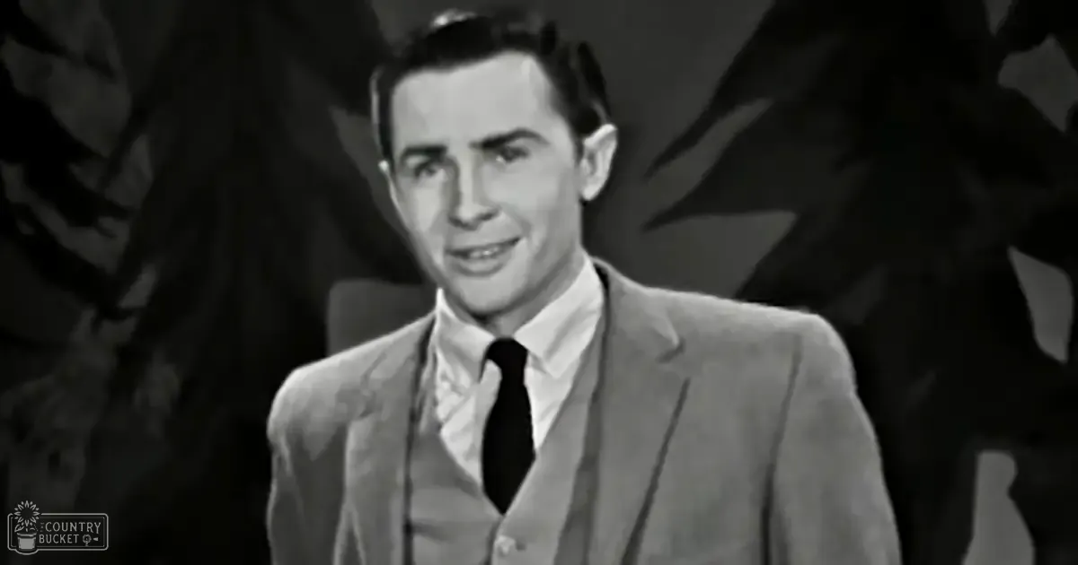 A young adult male in a three-piece suit stands on a stage-like setting with dark decorative elements behind him, suggesting an artistic or theatrical performance environment.