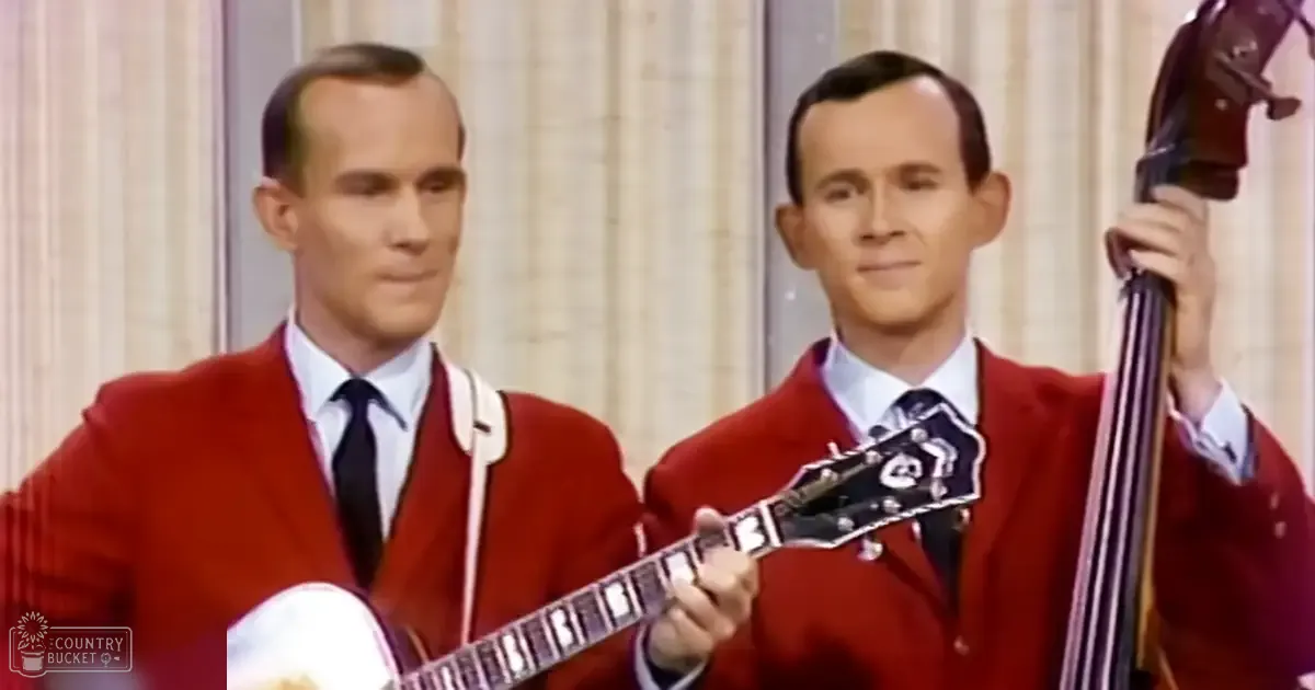 Two adult male singers in red blazers perform with a banjo and double bass against vertically striped beige curtains under bright studio lighting.