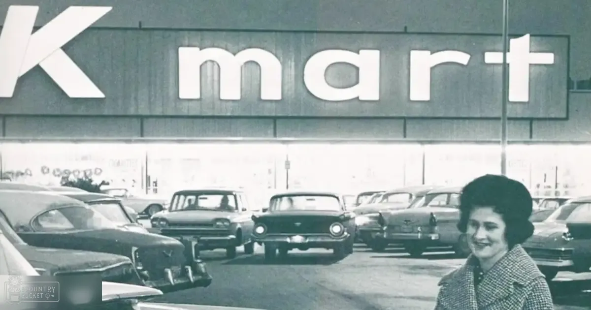 A woman in a checkered jacket stands before a Kmart store with cars from the 1950s visible in the parking lot, surrounded by lush greenery suggesting spring or summer.