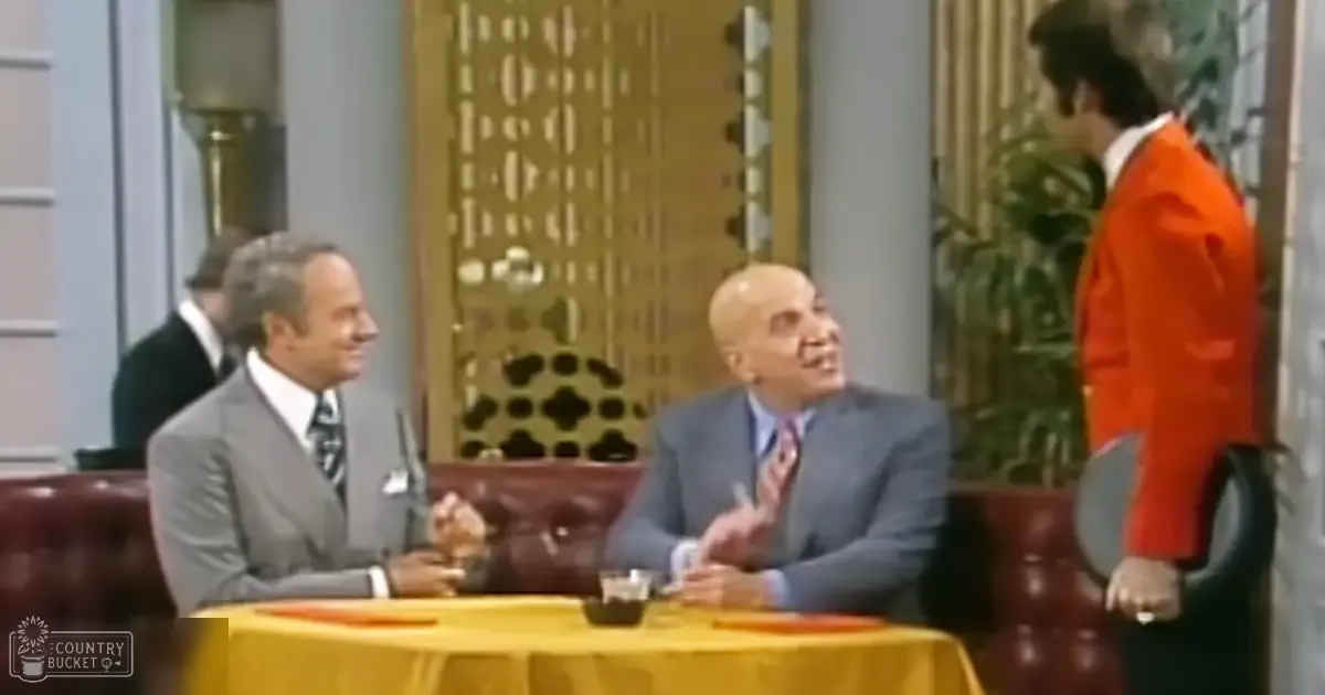 An indoor formal setting with a round yellow table, two red leather chairs, and decorative elements like a gold door, columns, and greenery; four men in suits seated or standing around the table.