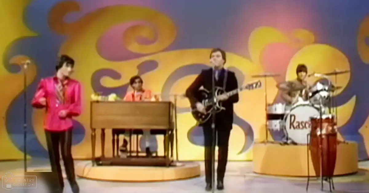 A mid-20th-century musical performance scene on a vibrant stage with swirling yellow and purple backdrop, with young adult male performers in suits and a grand piano.