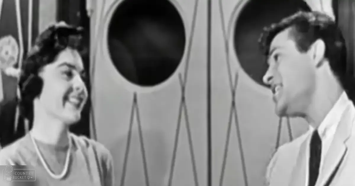 A mid-20th-century indoor scene with two young adults in light clothing, possibly a film or TV set, with subtle decor and natural daylight.