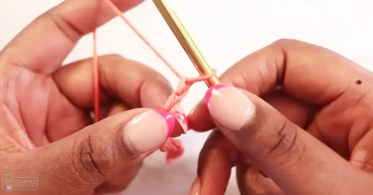 Close-up of hands knitting with pink yarn, golden needle, indoor setting, plain white background.
