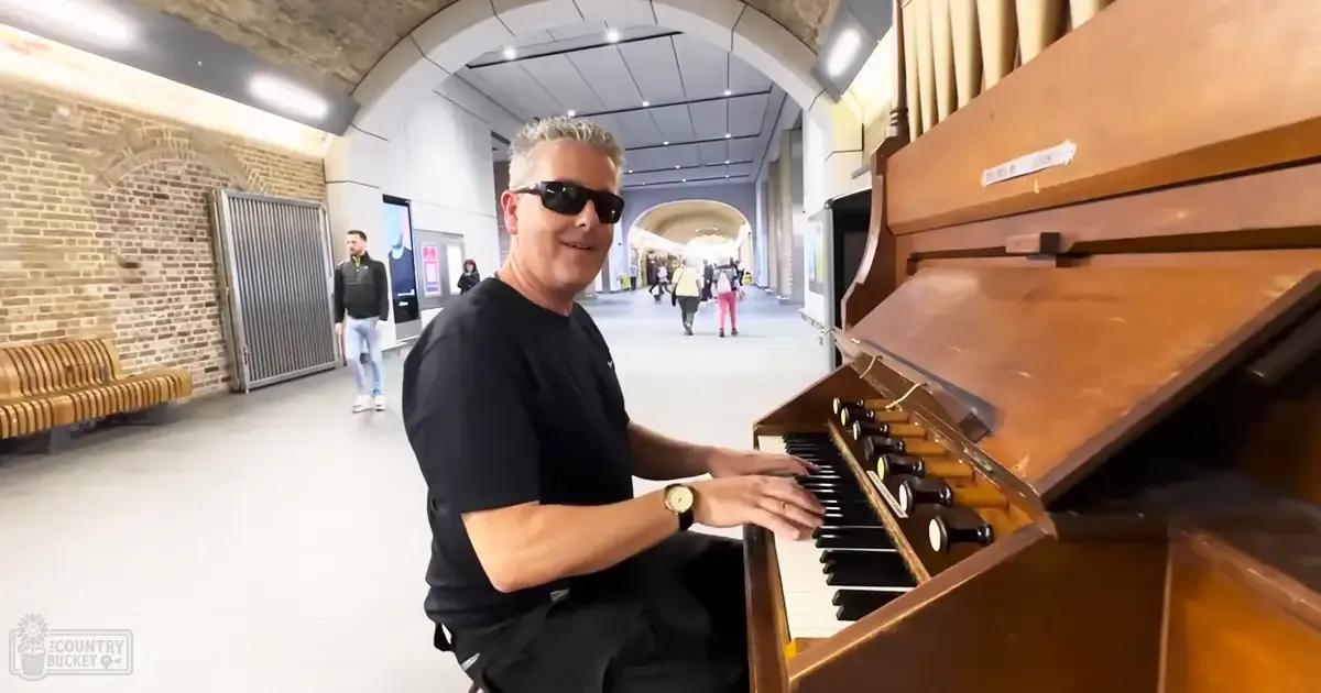 A middle-aged male playing an organ in a train station, surrounded by other passengers and signs.