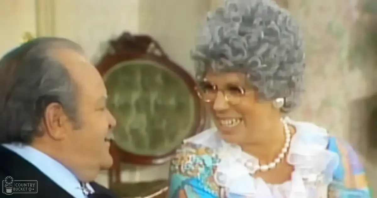 An older adult woman and a middle-aged man dressed in vintage attire stand indoors with a green upholstered chair and wallpapered background suggesting a mid-20th century home setting.