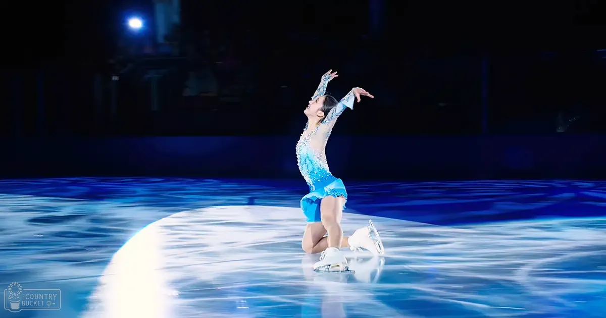A young adult female figure skater in a blue satin-like leotard with intricate designs and sequins, standing on an indoor ice rink at night.