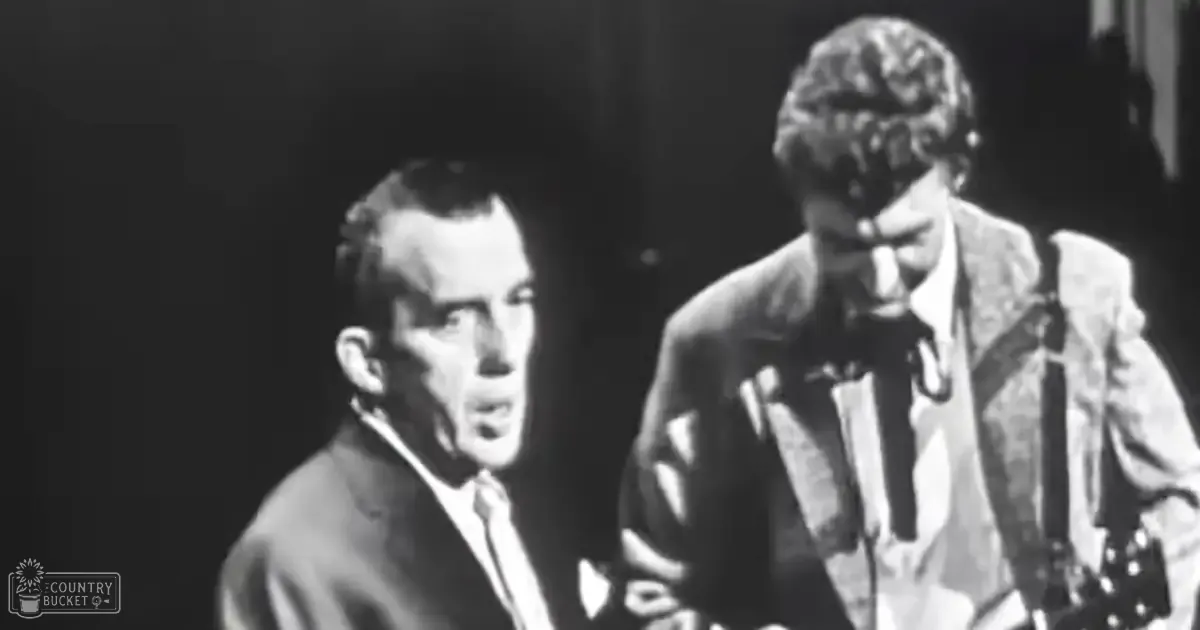 A musical performance scene, possibly from a concert or recording studio with two adult male performers dressed formally; one wearing a dark suit jacket over a white shirt and tie, the other in a light-colored textured suit jacket over a darker shirt or vest. Microphone stand visible behind them.