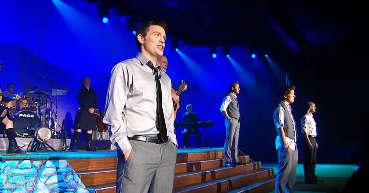 A young adult male singer in a light gray dress shirt and dark gray trousers performs on an indoor stage with dramatic blue lighting under wooden steps.