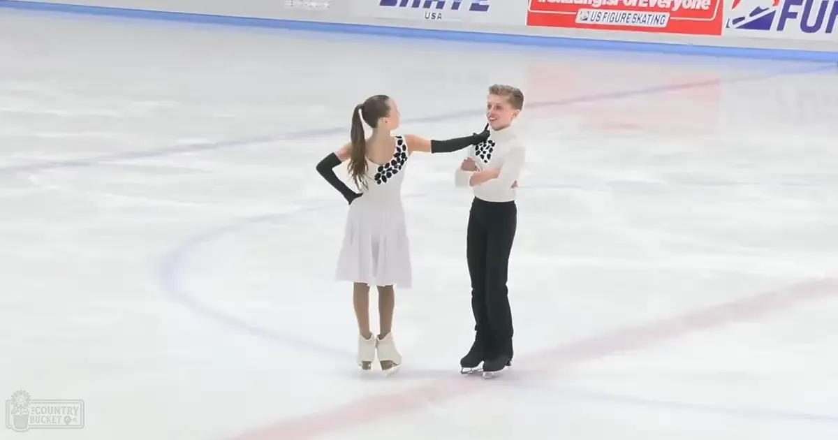 A young adult female and male figure skating pair in white dresses with black floral patterns and solid white tops, respectively, performing at an indoor ice rink under artificial lighting.