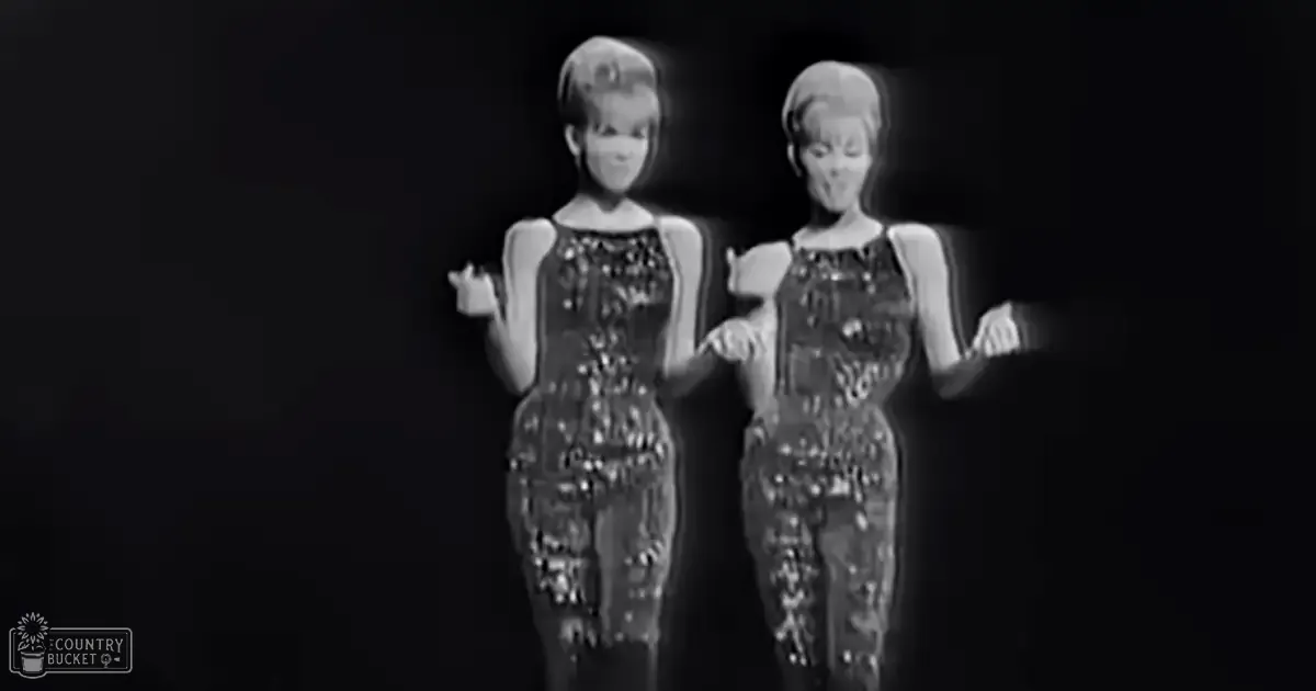 Two female performers in a vintage entertainment show, wearing sleeveless black-and-white beaded dresses with a scoop neckline and minimal lighting focused on them against a dark stage background.