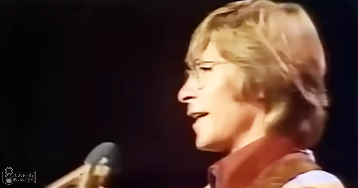 A young adult male singer performing on stage, wearing a brown 70s-style vest over a white shirt with short sleeves and collar under dimly lit stage lighting.
