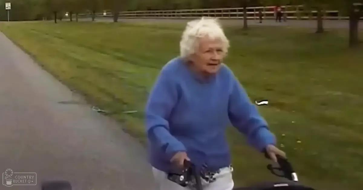 An older adult woman wearing a blue long-sleeved sweater and white pants is using a mobility scooter in a park setting with green grass, trees, and paved paths. The season appears to be spring or summer.