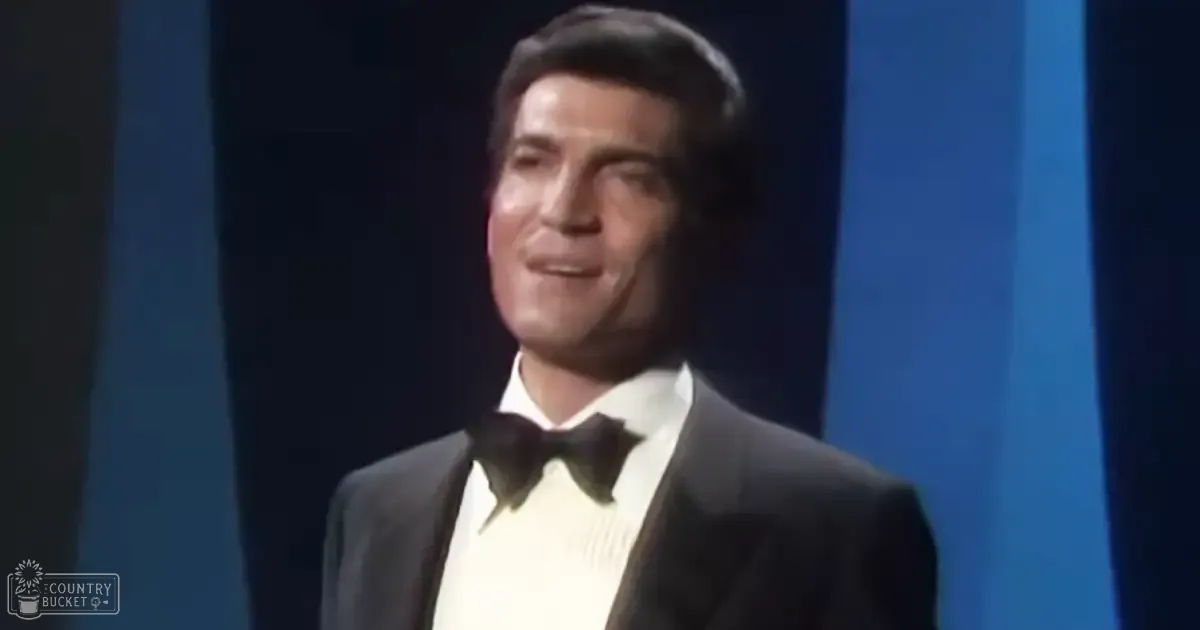 A middle-aged man performing on a theater stage wearing a classic black tuxedo with white dress shirt and black bow tie, set against dark curtains with vertical light strips.
