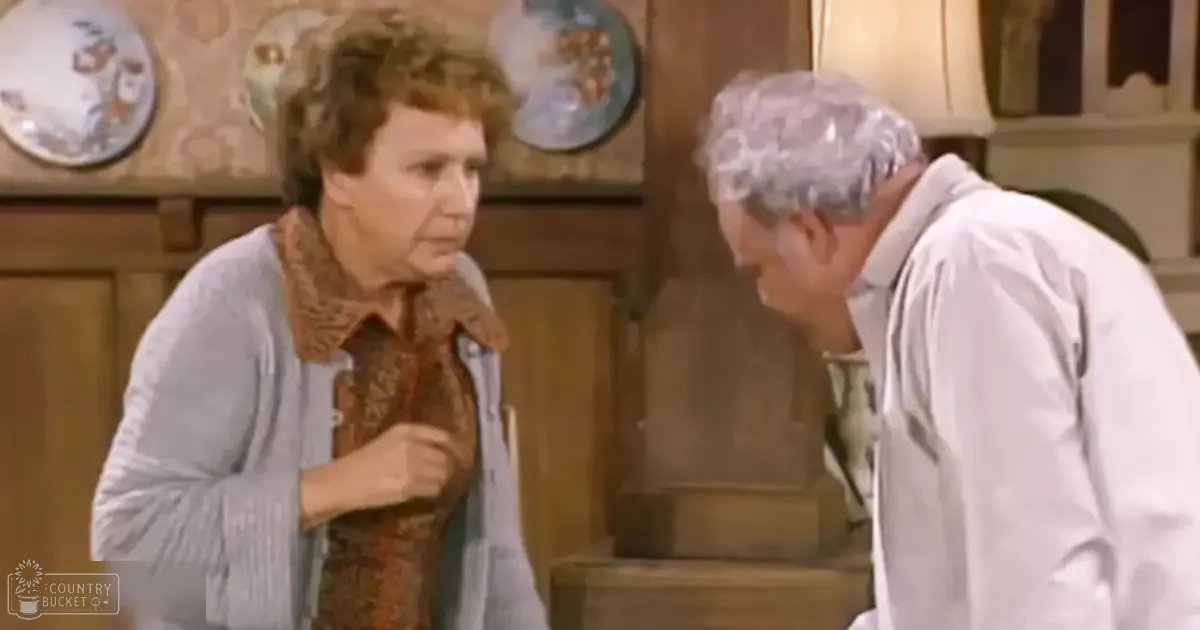 An older adult woman wearing a light blue striped cardigan and brown patterned blouse stands in a kitchen setting with wooden cabinets, decorative plates, and natural lighting.