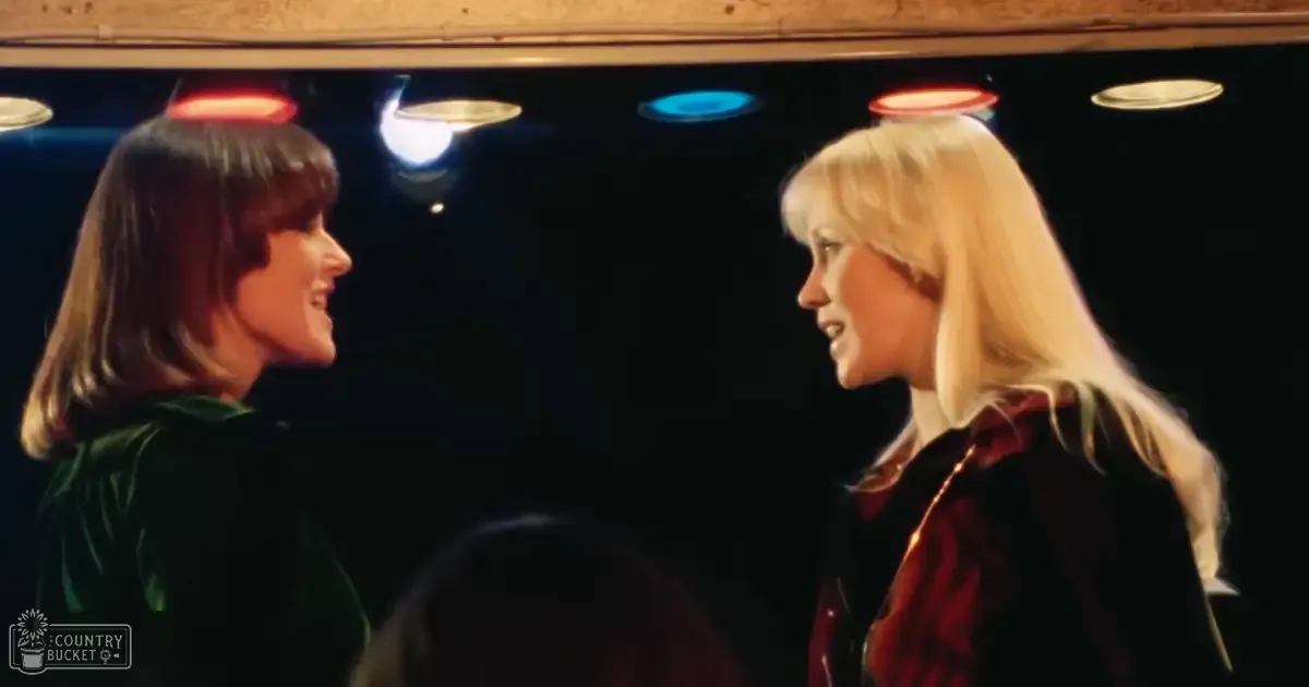 Two adult females in a theatrical setting, wearing dark green and red velvet garments with high collars, stage lighting with red and blue hues in the background.