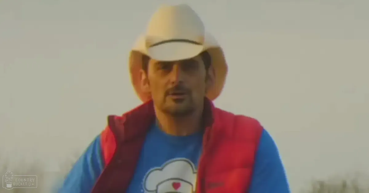 Middle-aged adult male wearing a white cowboy hat with dark band, blue graphic t-shirt with a smiling face and pink heart, red vest or jacket outdoors under an overcast sky.