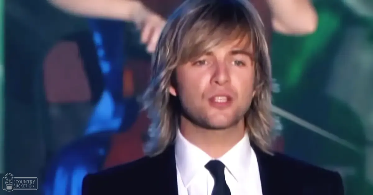 A young adult male wearing a black suit jacket, white dress shirt, and dark tie with classic knot style, standing indoors under stage-like lighting at an event or performance.