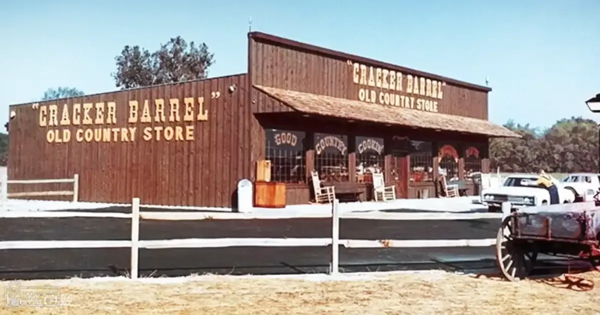 A Cracker Barrel Old Country Store in an outdoor rural setting with wooden storefront, white picket fence, rocking chairs, vintage car, and wagon.