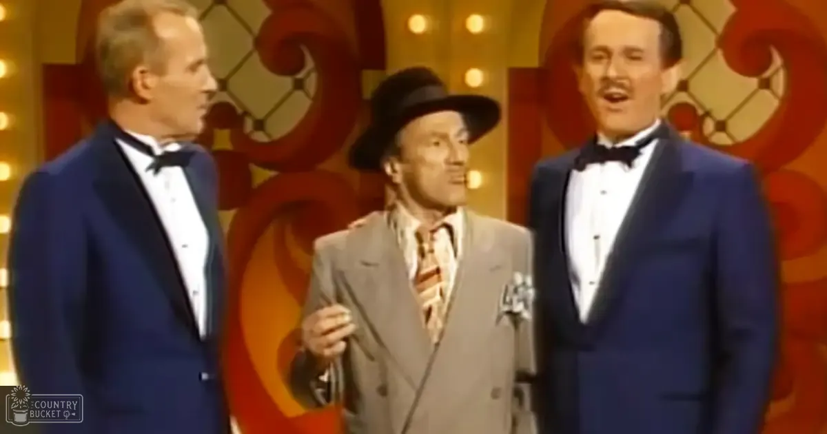 A group performance scene indoors with three adult male performers in formal attire against a backdrop of festive red and gold swirls under stage lighting.