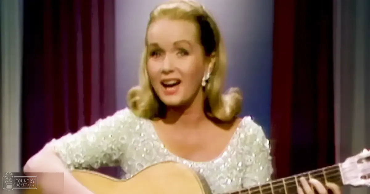 Adult female wearing a sparkly, sequined top with long sleeves and low neckline, possibly mid-20th century style, holding a guitar indoors on what appears to be a stage or studio. Text in bottom left corner reads 'Madly Odd.'