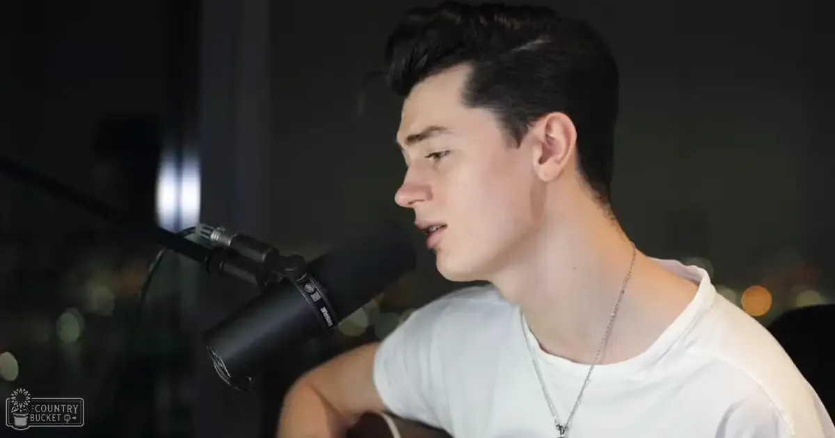 A young adult male in a relaxed white t-shirt, holding a Shure microphone, singing indoors under soft evening lighting.